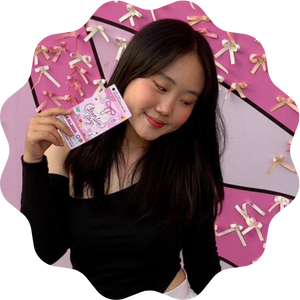 A young woman with long dark hair holding a pink card and looking to the side with a smile, standing in front of a pink backdrop decorated with bows.