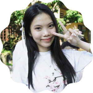 A young woman smiling and making a peace sign with one hand, wearing a white shirt with a floral design. She is outdoors with greenery and sunlight in the background.