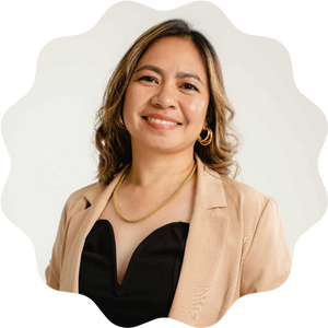A woman smiling at the camera, wearing a tan blazer over a black top, with shoulder-length wavy hair and gold hoop earrings, photographed against a light background.