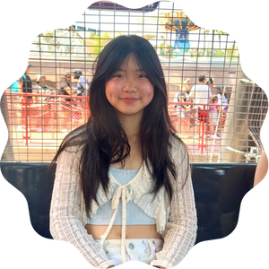 A young woman with long dark hair smiling softly while sitting in an amusement park ride, wearing a white knit cardigan over a light blue top. A metal gate and crowd are visible in the background.