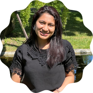 A woman smiling outdoors while sitting near a small pond, wearing a black blouse with lace details. Greenery and sunlight are visible in the background.