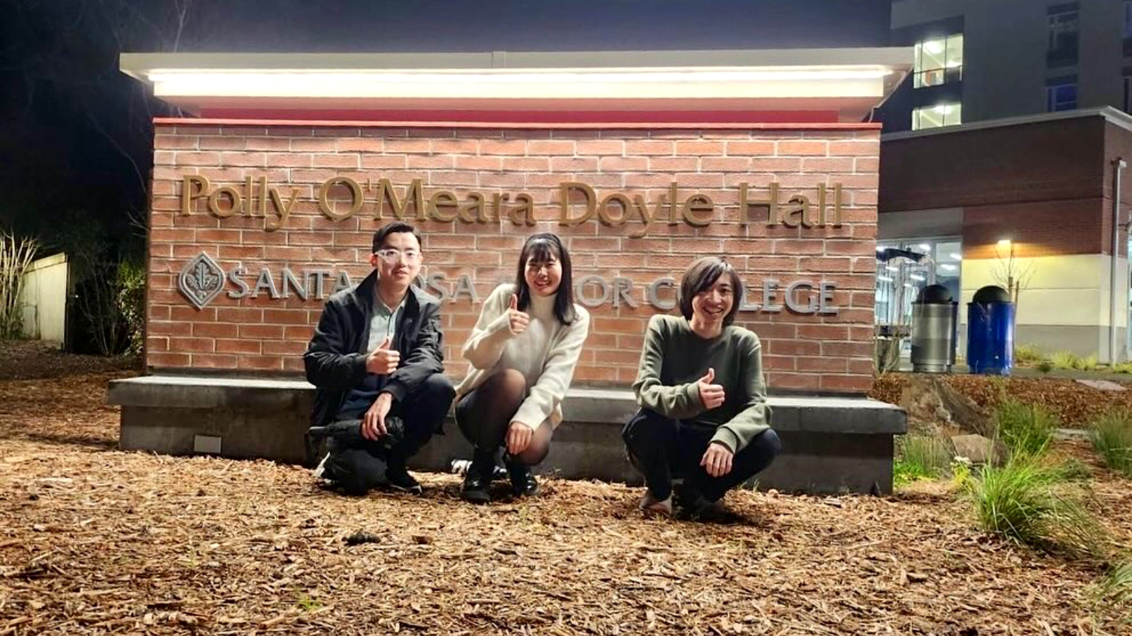 Three students posing in front of the illuminated Polly O’Meara Doyle Hall sign at night.