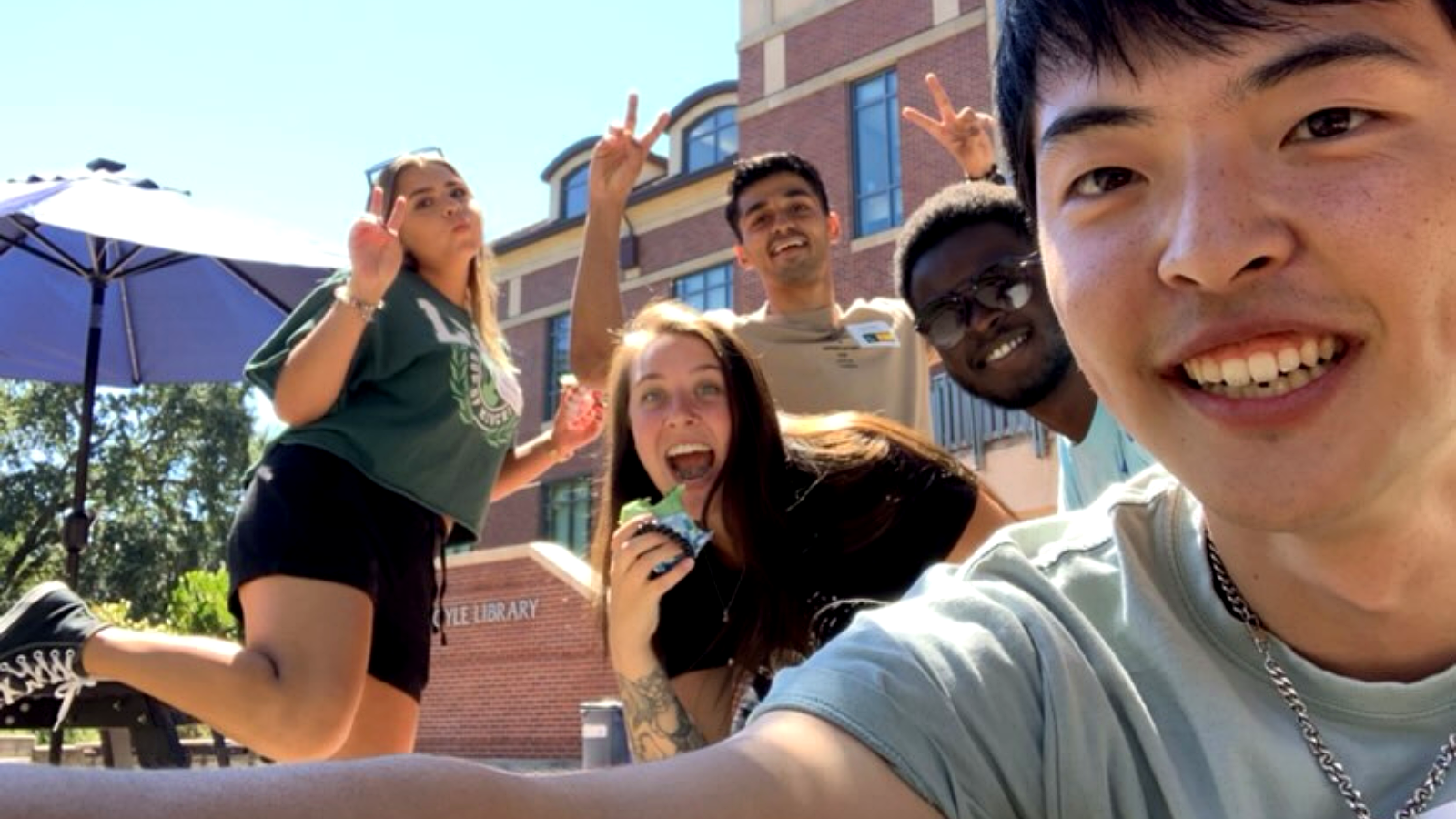 A group of students taking a cheerful outdoor selfie in front of Doyle Library, making peace signs and smiling.