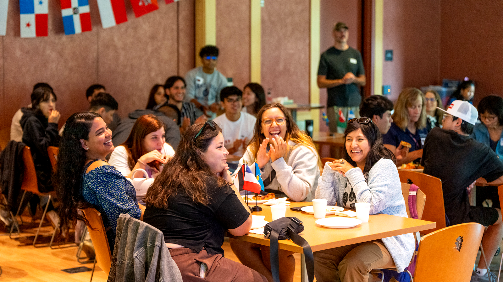 Students gathered indoors at an SRJC orientation event, smiling and talking around large tables with small international flags.