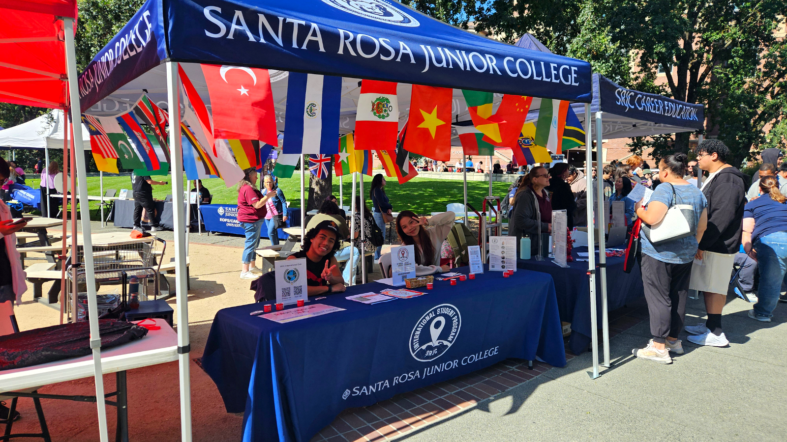 Students tabling at an outdoor SRJC event under a canopy decorated with international flags.