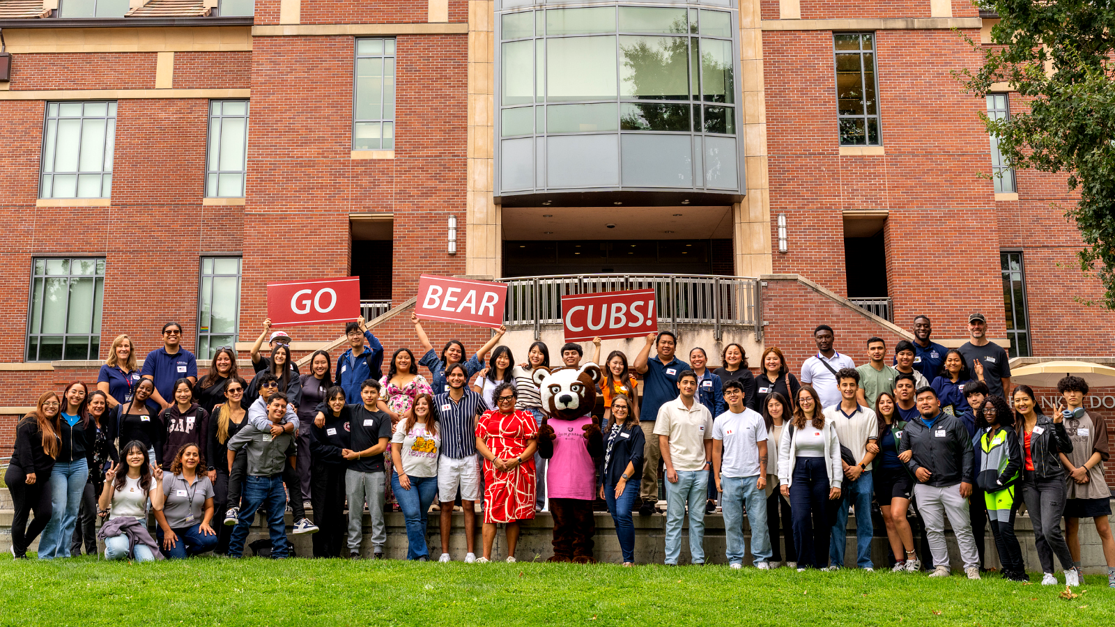 A large group of international students and staff posing in front of Doyle Library holding signs that say GO BEAR CUBS.